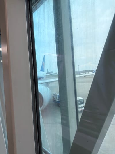 A view from inside an airport jet bridge shows an airplane parked on the tarmac. The tail of the airplane, featuring a blue and orange logo, is visible. A service vehicle is parked near the airplane. The image is taken from inside a jet bridge, looking out through the window at the airport scene. The location is Ota, Japan, likely at Haneda Airport.