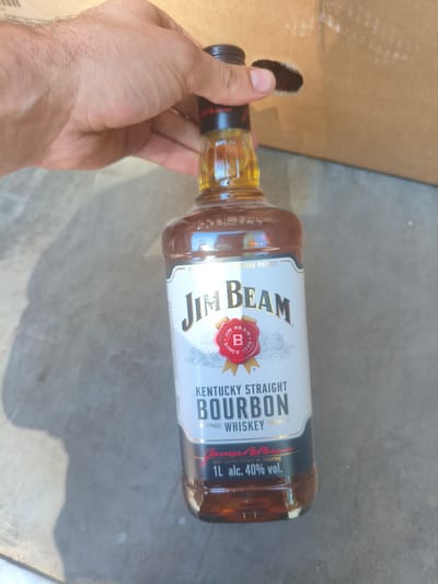 A hand holds a partially opened bottle of Jim Beam Kentucky Straight Bourbon Whiskey. The bottle is clear glass and filled with a dark amber liquid. The label is white with red and black lettering. The hand is holding the bottle near the top, with the thumb and index finger gripping the neck. The background is a textured gray surface, possibly concrete, with a shadow cast from the bottle. A cardboard box is visible in the upper right corner of the image. There is no information in the image to suggest it is located in Alchevsk, Ukraine.
