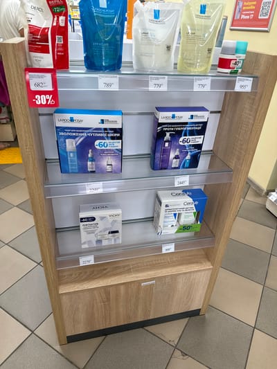 This is a photograph of a retail display shelf in a store, likely a pharmacy or beauty store, in Sarny, Ukraine. The shelf unit is made of wood-grain laminate with clear glass shelves. On the shelves are various skincare products, primarily from the brands La Roche-Posay and Vichy, as well as a Cerave product.
The top shelf displays several large pouches of liquid products, including La Roche-Posay Effaclar and Lipikar. There are also smaller Vichy bottles with discount stickers showing "-40%" and "-20%". A price tag of "955" is visible on this shelf.
The second shelf holds a La Roche-Posay set for sensitive skin, with a price tag of "2299".
The third shelf features a La Roche-Posay set for hyperpigmentation, with a price tag of "2445".
The bottom shelf displays a Vichy set for lifting and a Cerave set, with price tags of "2279" and "909" respectively. A red discount tag showing "-30%" and a price of "682" is attached to the wooden support structure on the left side of the display.
The floor is tiled with a pattern of beige and grey squares. The overall lighting is bright, suggesting an indoor commercial environment. No people are visible in the frame.