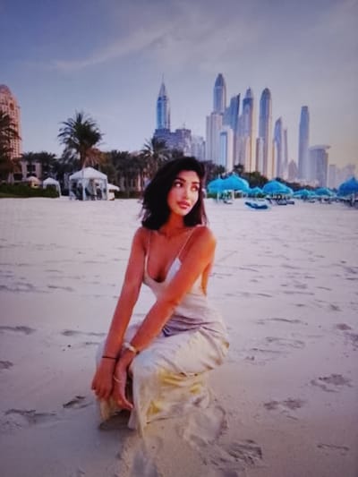 A woman with long dark hair is kneeling on a sandy beach, wearing a light-colored dress. She is looking to her left. In the background, there are palm trees, several buildings, and blue umbrellas on the beach. The sky is visible above, with some clouds. The location is the city of Prisel'e, Russia.