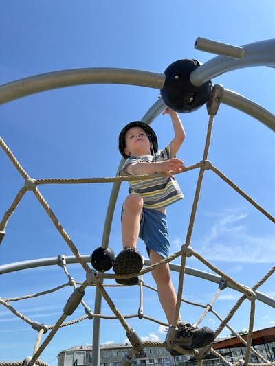 A young boy wearing a bucket hat, a striped t-shirt, and blue shorts is climbing on a dome-shaped climbing structure made of ropes and metal bars. He is positioned on the structure, with his right hand reaching up towards a black spherical joint and his left hand gripping a rope. The boy's legs are bent, and his feet are on different ropes. The climbing structure is set against a clear blue sky with a few wispy clouds. In the background, below the climbing structure, residential buildings are visible, suggesting an urban or semi-urban environment. The scene appears to be an outdoor playground.
