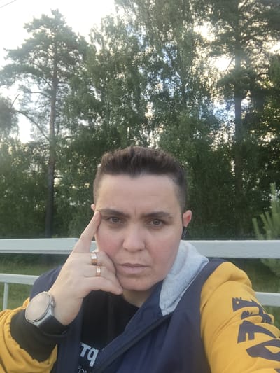 A woman with short, dark hair and a serious expression is taking a selfie outdoors. She is wearing a dark blue jacket with yellow sleeves and a grey hood, and a black t-shirt. She has two rings on her right hand, which is resting on her cheek. A silver smartwatch is visible on her left wrist. Behind her, there are tall green trees and a white railing. The image appears to have been taken in the village of Tsibino, Russia.