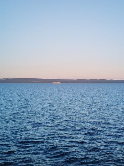 The image captures a serene seascape at dusk or dawn, with the sky transitioning from a pale blue at the top to a soft pinkish hue closer to the horizon. The vast expanse of the water is a deep blue, with gentle ripples reflecting the diffused light. In the distance, a long, tree-covered shoreline stretches across the frame. A white passenger ferry or large boat is visible on the water, approximately in the middle of the frame, with a smaller vessel to its right. The overall scene conveys a sense of tranquility and natural beauty. The image is a photograph taken in Kakkarovo, Russia.