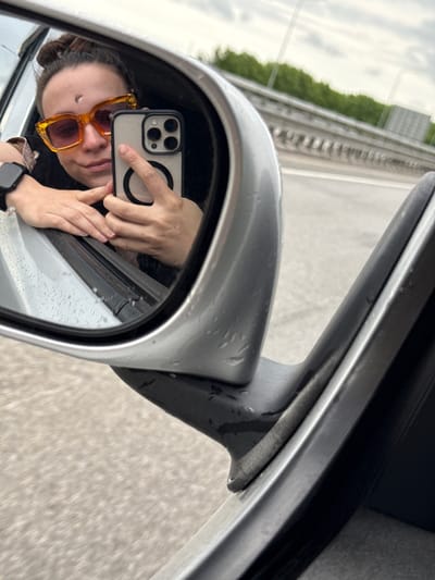 A person is taking a selfie in a car's side mirror. The person has sunglasses and a watch. The environment appears to be a road with a highway divider in the background. Based on the prompt, the location is assumed to be in the city of Shali, Russia.