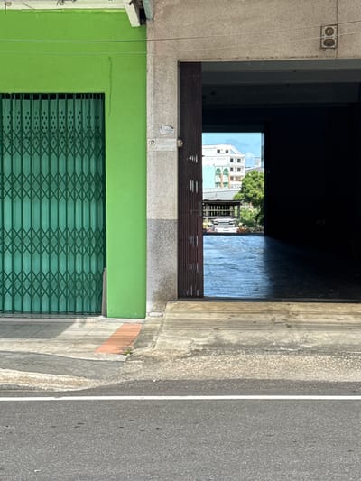 The image depicts a street scene in Ranong, Thailand, focusing on the entrance to a building and its exterior. On the left is a bright green building with a closed, dark green, metal accordion gate covering its storefront. The gate has a diamond pattern. Adjacent to the green building, to its right, is a concrete structure with a rough, mottled grey texture. This structure has an open doorway revealing a dark interior space, partially illuminated by natural light from beyond. Through the open doorway, a sunlit scene of a courtyard or pathway is visible, with a reflective, wet-looking surface. In the distance, a light-colored building with green accents and a white rooftop structure can be seen, along with trees. The ground in front of the buildings consists of concrete sidewalks and a section of asphalt road with a white stripe. The overall impression is one of an urban environment, possibly a commercial area, captured on a bright day.