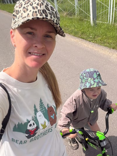 In the city of Kul'dur, Russia, a woman wearing a leopard-print baseball cap and a white t-shirt with a "Bear Brothers" graphic is taking a selfie. She is standing to the left, with her face and upper body visible. To her right, a young child, wearing a green-patterned bucket hat and a brown jacket, is seated on a black and green bicycle. The child is looking down and to the right. Both subjects are on an asphalt road. In the background, there is green grass and a white fence. The lighting suggests it is daytime.