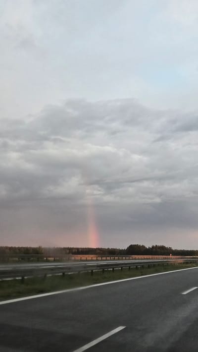 The image depicts a highway scene under a cloudy sky. A faint rainbow is visible in the distance, appearing as a vertical streak of pink and red light. The road is wet, suggesting recent rain. A metal guardrail separates the highway from a grassy embankment and a dense line of trees in the background. The sky is overcast with varying shades of gray and white clouds. There are no people or vehicles clearly visible in the image. The location is identified as Rosin, Poland.