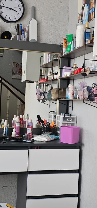 The image shows a shelf with various personal care items and accessories. The shelf is dark-colored and appears to be made of wood or a similar material. On the shelf, there are bottles of lotions and sprays, makeup brushes, hair clips, sunglasses, and a clock. A framed picture of a young girl is also visible. To the right of the shelf, there are several white rectangular panels, possibly part of a larger furniture piece or decorative elements. The overall scene suggests a personal storage area, likely in a bedroom or bathroom. The image is taken from a slightly elevated angle, and the lighting suggests it was taken indoors during the daytime. There is no indication of weather or specific time of day beyond daylight. No text is clearly legible in the image.