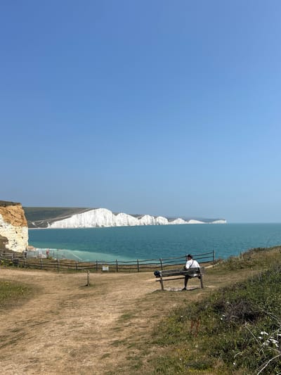 The image depicts a scenic coastal view of the Seven Sisters cliffs in Seaford, United Kingdom. In the foreground, a wooden bench sits on dry, brown grass, facing the sea. A person is seated on the bench, facing away from the camera, wearing a white t-shirt and dark shorts, with a black hat. A wooden fence runs along the edge of the grassy area, separating it from the sea. The sea is a vibrant turquoise color, and in the distance, the iconic white chalk cliffs of the Seven Sisters stretch across the horizon under a clear blue sky. The overall scene is one of natural beauty and tranquility.