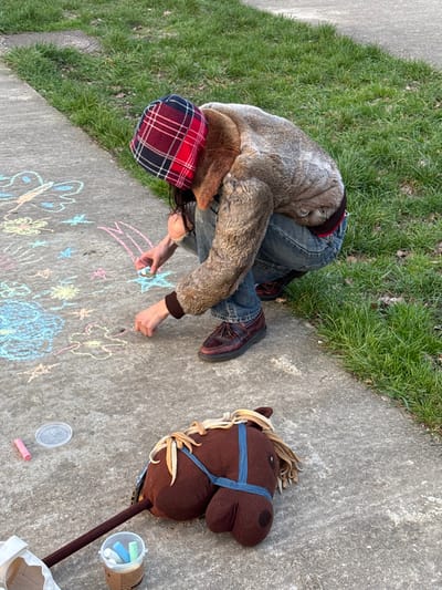 This image captures an individual crouching on a concrete sidewalk in Saint-Mandé, France, engaged in drawing with chalk. The person is wearing a red and black plaid bucket hat, a light brown fur-textured jacket with dark brown cuffs, blue denim jeans, and dark red-brown tassel loafers. Their right hand holds a white chalk stick, actively adding to drawings on the pavement. The sidewalk features multiple colorful chalk drawings, including a light blue butterfly, yellow flowers, pink and blue stars, and other abstract shapes. Resting on the sidewalk to the lower right is a brown plush hobby horse with a dark brown stick, a tan mane, and a blue strap across its back. Near the hobby horse, a clear plastic container holds several assorted colored chalk sticks, and a single pink chalk stick lies beside a transparent plastic lid. A portion of a light-colored canvas bag is visible at the bottom left. The concrete sidewalk is bordered by a strip of green grass on the right side of the frame. The scene is illuminated by natural daylight.