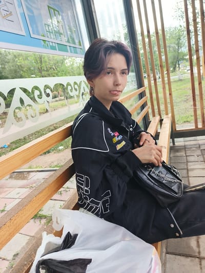 A young person with dyed purple and black hair is sitting on a wooden bench at a bus stop in Arkalyk, Kazakhstan. They are wearing a black jacket with white detailing and patches, and black pants. A black purse with a silver chain strap is on their lap. In front of them are two plastic bags, one white and one black. The bus stop has a glass enclosure with a wooden frame and decorative patterned glass. In the background, there are trees and a street.