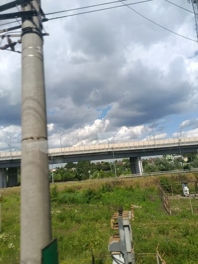 The image shows a view of an elevated road overpass with a concrete pillar in the foreground, on the outskirts of Kupavna, Russia. The pillar is a light gray, cylindrical structure, partially obscuring the view of the surrounding environment. Above the overpass, a cloudy sky dominates the upper portion of the frame. The overpass itself is a multi-lane structure with safety barriers and lighting. Below the overpass, a landscape of green vegetation, trees, and some buildings can be seen. In the foreground, some electrical equipment is also visible, suggesting a location near a transportation route or utility infrastructure. The overall scene depicts an urban or suburban setting with infrastructure and natural elements.