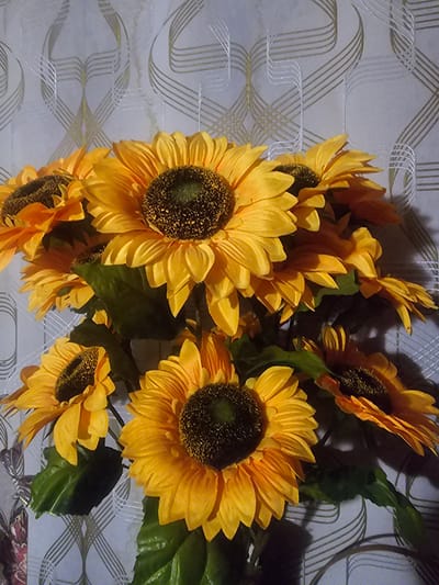 A bouquet of artificial sunflowers is displayed against a patterned wallpaper. The sunflowers are a vibrant yellow with dark centers and green leaves. The wallpaper features a repeating pattern of interwoven, wavy lines in shades of gray and gold. The lighting in the image is somewhat dim, casting shadows on the flowers and wallpaper. The scene is likely an indoor setting, possibly a home or a commercial establishment, and there is no indication of a specific location in Taezhnoye, Russia.
