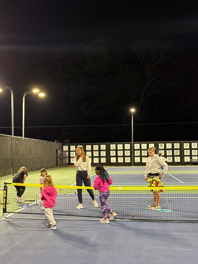 Five people stand on a tennis court at night. A yellow net separates them. Overhead lights illuminate the court, and behind them is a dark background with trees and light poles. The people appear to be leaning or stretching over the net. Tennis balls are scattered around the players. There is a scoreboard in the background.