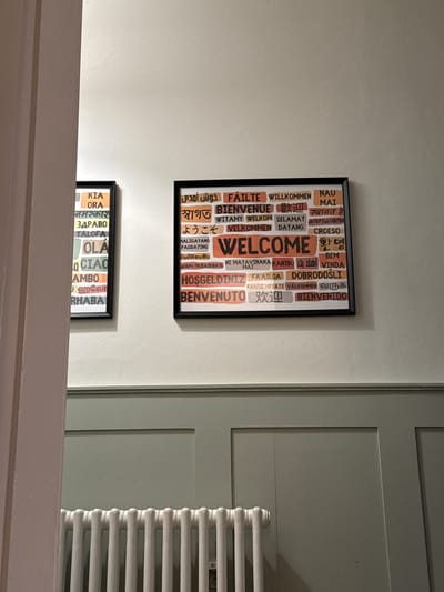 The image shows a section of a wall, featuring two framed "Welcome" prints in different languages. The upper print is larger and includes various translations of "Welcome", such as "Fáilte", "Willkommen", and "Bienvenue". The lower print is smaller and features translations such as "Kia Ora" and "Ciao". Adjacent to the wall, to the right, is a white radiator and a gray-painted wooden wall paneling. The scene appears to be indoors, likely in a home or building within Windermere, United Kingdom.