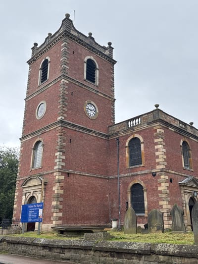 The image displays an exterior view of a historic church building, identified by a visible sign as St John the Baptist, Knutsford Parish Church. The scene is captured on an overcast day, providing diffused lighting.
The church is constructed predominantly from red brick, contrasted with lighter, possibly sandstone, quoins and decorative bands that highlight its architectural features. A prominent multi-tiered tower stands to the left, featuring arched windows with dark panes and at least two circular clock faces, one near the top and another lower down, both displaying Roman numerals. The tower's top is adorned with ornamental finials.
The main body of the church extends to the right, also showcasing arched windows and a stone balustrade along its roofline. In the foreground to the right, a grassy area contains several dark, weathered grave markers or headstones, bordered by a rough-hewn stone wall. Green foliage is visible at the bottom right corner of the frame.
Near an entrance on the church's side, a blue sign is clearly legible, stating: "St John the Baptist", "KNUTSFORD PARISH CHURCH". Further text details service times, including "Sun 8am Holy Communion", "9:30am Morning Worship", "6:30pm Evening", with additional, partially visible entries for other weekdays like "Tue", "Wed", "Fri", and "Sat". No specific activity or people are depicted; the image is a static architectural and landscape shot.