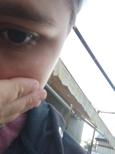 An extreme close-up selfie shows the left eye of a person. The eye is dark and the iris is barely visible. The person's hand is covering the lower part of their face, with the thumb and index finger obscuring the mouth and chin. The individual is wearing a dark hooded sweatshirt with a white logo on the left side of the hood. In the background, a structure with a wooden beam and a series of white and yellow flags or prayer banners is visible. The sky is bright and overcast. The image is grainy and lacks detail, making it difficult to ascertain the precise setting. The scene appears to be outdoors, possibly in a rural or semi-urban area of Cîrpești, Moldova.