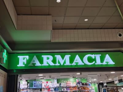 A brightly lit, green sign with white lettering that spells "FARMACIA" is prominently displayed above the entrance to a pharmacy. To the left of the sign, a neon green cross, a symbol commonly associated with pharmacies, is visible. Below the sign, the interior of the pharmacy is visible, showcasing rows of shelves stocked with various products, including medications and personal care items. Several cash registers, labeled "Caja 4" and "Caja 5," are visible, indicating customer service points. The ceiling above the pharmacy is tiled, and modern light fixtures are embedded in it. The overall scene suggests a well-organized and accessible pharmacy environment.