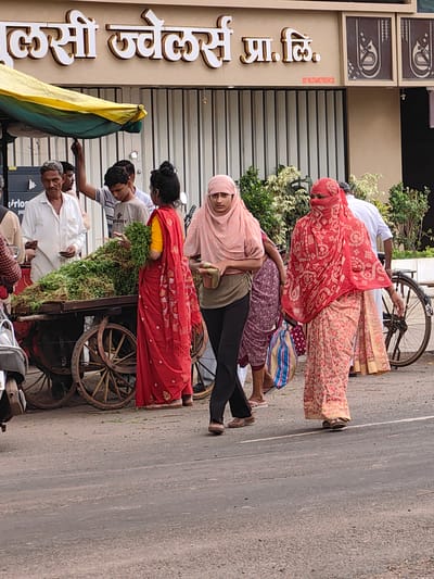In Datala, India, a street scene unfolds near a jewelry store. A woman in a vibrant red and pink floral sari, her face covered by a patterned red veil, walks towards the right, carrying a striped bag. Beside her, a woman in a pink hijab and a green top walks past a cart laden with green produce, dressed in a flowing red sari. Another woman in a dark patterned skirt and a maroon hijab stands behind her. In the background, several men are visible near a stall with a yellow and green awning, one man in a white shirt looking towards the cart. A bicycle with a large wheel stands to the right, partially obscured by the people and the cart. The street surface is asphalt, with a white line marking a lane. The building behind them features a sign in an Indian script and decorative panels.