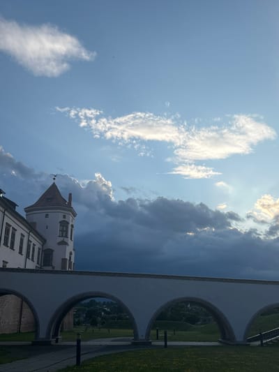 The image depicts a view of Mir Castle, located in the town of Mir, within the Karelichy District of Grodno Region, Belarus. The castle, a UNESCO World Heritage site, is partially visible on the left side of the frame, showcasing its distinctive white walls and a red-tiled tower with a weather vane. In the foreground, a white arched bridge or walkway, likely part of the castle's grounds or surrounding park, stretches across the lower portion of the image. Beyond the bridge, a green grassy area with scattered yellow flowers and a few light posts can be seen. The background is dominated by a dramatic sky filled with a mix of dark, stormy clouds and bright, sunlit clouds, suggesting a dynamic weather condition. The overall scene captures a historical landmark against a striking natural backdrop.