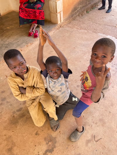 This outdoor, high-angle shot captures three young children interacting playfully on a light brown, dusty ground in Rahwol Chom, Nigeria, under bright daytime lighting.
In the foreground, a child lies on their back, wearing a light brown, possibly traditional, long-sleeved garment. Their arms are crossed, and they look up at the camera with a playful smile and their tongue sticking out. Above them, another child is also lying on their back, wearing a light-colored camouflage-patterned t-shirt and dark pants. This child looks directly at the camera with a wide smile and extends one arm towards the left. Both children have short, dark hair and joyful expressions.
The third child stands behind and above the other two, facing the camera. They wear a grey and red short-sleeved t-shirt with a graphic design, blue shorts, and a grey clog on one foot. This child makes a peace sign with their right hand, smiling broadly. Some white residue is visible on their nose and cheek.
Partial views of two adults are also included in the frame. On the far left, a person's lower legs and feet, clad in red sandals and a vibrant black garment with red and gold patterns, are visible. In the upper left corner, another person's legs and black shoes are seen standing near some small plants. The children appear happy and engaged with the camera, contributing to the candid and joyful atmosphere of the scene.