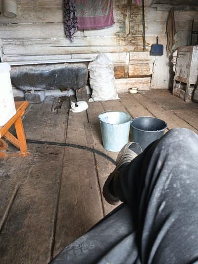 The image depicts the interior of what appears to be a rural outbuilding or shed, possibly in or near Uvat, Russia. The floor is made of rough wooden planks, showing signs of wear and dirt. In the foreground, a person's legs, clad in dark pants and gray shoes, are visible, suggesting the viewpoint is from someone seated or resting.

The room contains several objects. A white cat with black markings is lying on the floor near a large, rough-hewn stone trough. To the right of the cat are two buckets: one light blue and metal, the other black and plastic. A large sack of white material, possibly grain or feed, stands upright in the middle ground. The walls are constructed of weathered, horizontal wooden planks. Various items are hung on the wall, including a patterned cloth or towel, a dustpan, and another piece of fabric. In the background, there is a rustic, metal stove or oven-like structure. The overall impression is one of a utilitarian, somewhat unkempt, working space.