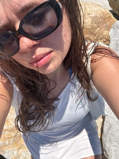 The image is a close-up, selfie-style shot of a young woman wearing black sunglasses and a white dress. She has brown hair and her skin appears tanned. There are some blemishes visible on her cheek. The background consists of rocks and what appears to be a sandy or rocky shoreline. The lighting suggests it is daytime and sunny. The image does not provide specific visual cues to identify the location as Nea Irakleitsa, Greece.