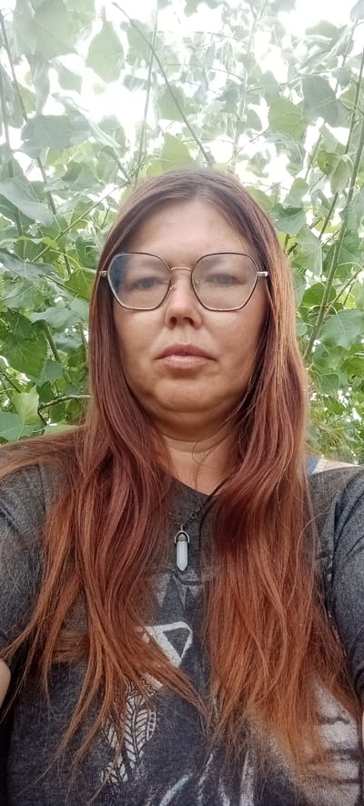 A selfie of a woman with long, auburn hair and glasses is taken outdoors. The woman is wearing a dark grey t-shirt with a white graphic print and a necklace with a white, pointed pendant. She is positioned centrally in the frame, with lush green foliage providing a blurred background. The location is Январцев ауылдық округі, Kazakhstan.