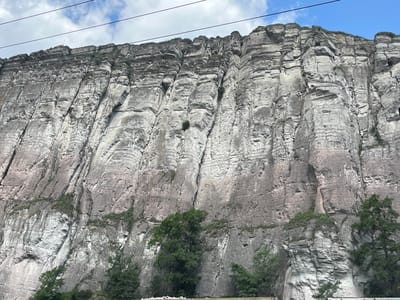 The image shows a massive, sheer cliff face, likely made of sedimentary rock, with distinct horizontal layering and vertical fissures. The rock is predominantly gray with streaks of pinkish-brown. Sparse vegetation, consisting of small bushes and tufts of grass, clings to ledges and crevices near the top and in some sections of the cliff face. At the base of the cliff, a line of trees with dark green foliage is visible, suggesting a natural setting. The sky above is partially cloudy, with patches of blue sky visible between white and gray clouds. Several dark, thin power lines or cables stretch horizontally across the upper portion of the image, indicating the presence of human infrastructure. The overall impression is one of a grand and rugged natural landscape, possibly a roadside view given the presence of the cables. There is no visible text in the image. The lighting suggests it is daytime, likely with a mix of sun and cloud cover.
