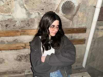 A person with dark hair and glasses is upside down in a room with a concrete floor. The person is wearing a grey hooded jacket, a white shirt, and blue jeans. A white pipe runs along the left side of the image. Wooden planks are attached to the floor. A metal grate is visible near the bottom left of the image. The location is unknown, and there is no confirmation that this image was taken in Agarak, Armenia.