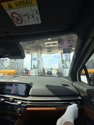 Inside a car approaching a toll booth, the dashboard dominates the view. A navigation app is visible on a phone mounted on the dash. Outside the car, toll booths stand in rows under a bright sky. A black car is stopped ahead, awaiting passage. Signs indicate toll fees. Part of a white sock is visible in the upper right corner of the car. A warning sticker on the driver's side regarding airbag deployment can be seen on the visor.