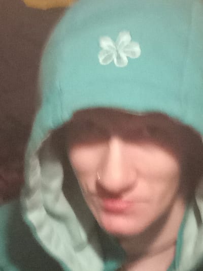 The image shows a close-up of a person's face. The person is wearing a turquoise hooded sweatshirt with a white flower design on the hood. Their face is obscured by the hood, only their nose and mouth are visible. They are wearing a nose ring. The person appears to be looking down and their expression is neutral. The background is out of focus and appears to be a brown wall. The image was likely taken indoors. The latitude and longitude provided correspond to a location in the city of Kitchener, Canada. However, the image itself does not provide any information about the location.