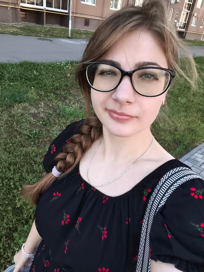 A young woman with light brown hair styled in a braid and wearing black-rimmed glasses takes a selfie outdoors. She is dressed in a black off-the-shoulder top adorned with red floral patterns and has a silver chain necklace. Her right arm is extended, holding what appears to be a light blue bag or jacket. A watch is visible on her left wrist. The background features a paved walkway, a grassy area with scattered white flowers, and a multi-story brown building with numerous windows. The setting is identifiable as Novaya Usman, Russia, based on the architectural style of the building and the overall environment.