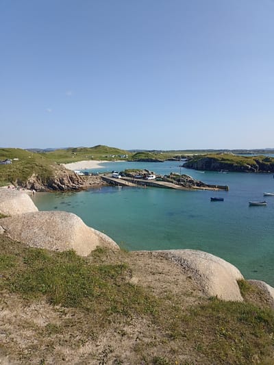The image depicts a scenic view of a harbor and beach area in Carnboy, Ireland, under a clear blue sky. The foreground is dominated by large, light-colored boulders covered in sparse green grass and some scattered debris. In the mid-ground, a calm, turquoise body of water laps against a rocky shoreline and a concrete pier. Several small boats are moored in the water near the pier, and some vehicles, including white vans and cars, are parked on the pier itself. A sandy beach stretches along the coastline, dotted with a few individuals. Beyond the beach, rolling green hills rise, with more buildings visible in the distance. The overall scene is one of a peaceful, sunny day at a coastal location.