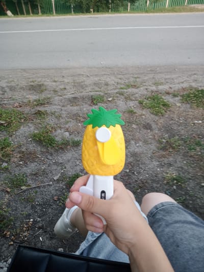 A person's hand holds a yellow pineapple-shaped fan in front of a blurred background of a road and greenery. The fan has green leaves on top and a white button and mechanism. The person is wearing denim shorts and white sneakers. The scene appears to be outdoors, possibly near a street in Elizaveta, Moldova, given the context. The ground is covered in dirt and sparse vegetation.