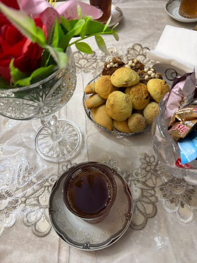 This is a still-life photograph taken indoors, likely on a table covered with a decorative cloth featuring floral embroidery. The scene suggests a relaxed or celebratory setting.
In the foreground, a clear glass teacup filled with dark liquid, presumably tea, sits on a white saucer with an ornate grey and brown pattern. To the left, a crystal vase holds a vibrant red artificial rose and green artificial leaves. Behind the vase, a clear glass pedestal dish supports a decorative glass piece.
Dominating the mid-ground is a crystal dish filled with small, golden-brown baked goods, possibly cookies or small cakes. Some of these appear to have nuts or dried fruit embedded in them. Next to this dish, a stack of folded white napkins is visible. To the right, a clear plastic container overflows with individually wrapped candies in colorful foil packaging, predominantly pink and gold.
The background is slightly blurred, but it appears to be an indoor environment. There are no people visible in the image. The lighting is soft, creating gentle highlights and shadows. The overall impression is one of an inviting spread of tea and sweets.
While the image depicts a scene consistent with a home or a café setting, there is no direct visual information that can place this specific scene within Balakənd, Azerbaijan. The objects and decorations are not culturally specific to that region.