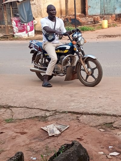 An adult male is seated on a dark blue and black "Qalink" brand motorcycle, stopped on a dirt shoulder adjacent to a paved road in Oko Town, Nigeria. The man, with a dark complexion and short hair, wears a white t-shirt, light-colored trousers, and black slides. A black shoulder bag with a white emblem is slung across his chest. He holds the motorcycle's handlebars, with his feet resting on the ground, and looks towards the viewer's left. The motorcycle features a prominent round headlight, a metallic engine guard, and a chrome exhaust. In the background, a light-colored building with a grey awning and a white poster is visible to the left, and a weathered building with a blue facade and a yellow jerrycan is to the right. The foreground shows reddish-brown dirt with cracks and sparse vegetation, where a crumpled piece of paper or cardboard lies flattened.