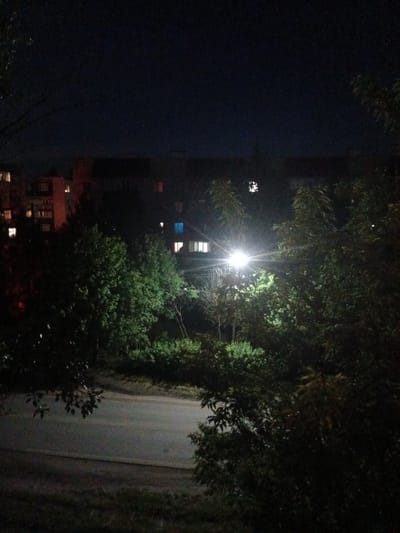 The image captures an outdoor scene at night in the city of Kuznetsk, Russia. The foreground is dominated by dark foliage and trees, with a paved road visible below and in the middle ground. A bright street lamp illuminates the area, casting a strong glare and creating starburst effects. In the background, an apartment building is visible, with several windows lit from within, suggesting occupied residential units. The sky is dark and clear. The scene appears to be a quiet residential street with vegetation and typical urban infrastructure.