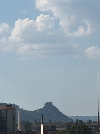 This media file captures a panoramic view of a cityscape against a backdrop of a mountain range and a cloudy sky. The composition is divided, with the left side dominated by soft, white clouds against a pale blue sky, suggesting a daytime scene. Small, dark specks, possibly birds, are visible against the clouds. The right side of the image reveals a more structured environment, characterized by a chain-link fence in the foreground. Beyond the fence, buildings with metallic siding and brown roofing are discernible, along with glimpses of trees and a distant mountain. The mountain itself is covered in lush greenery, with a distinct, rocky peak. In the far distance, faint outlines of what appear to be wind turbines can be observed, hinting at the presence of industrial infrastructure. Two white cylindrical structures, possibly industrial chimneys or ventilation shafts, are also visible in the mid-ground. The lighting suggests it is either morning or late afternoon, with the sun casting a soft glow. The overall atmosphere is one of tranquility, with the natural landscape meeting the urban environment. There is no visible text in the image. The context suggests the location is Singen (Hohentwiel), Germany, which aligns with the presence of a prominent mountain and a developed urban area.