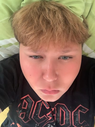 The image is a close-up, high-angle, selfie-style shot of a young person with light brown hair and fair skin. The person is wearing a black t-shirt with a red and white AC/DC logo. They are lying down on a green and white striped surface, possibly a bed or sofa. The background is a blurred green fabric, suggesting an indoor setting. The image does not contain any information about the city of Vejprty, Czechia.