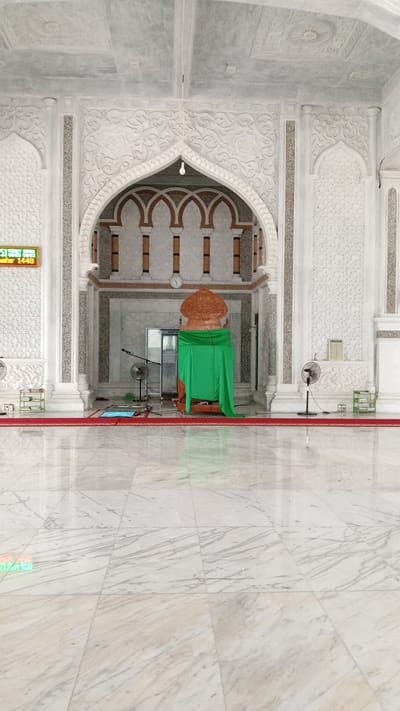 The image shows the interior of a mosque. A minbar, covered with a green cloth, is situated in the center of the frame. Two fans are positioned on either side of the minbar. A prayer mat is laid out in front of the minbar. The walls and ceiling are adorned with intricate white carvings. A digital clock displaying "1448" is visible on the left side of the image. The floor is made of polished white marble. The scene appears to be captured in the city of Beunyot, Indonesia.