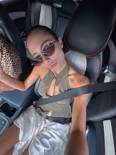 A person is reclining in the driver's seat of a car. The person is wearing sunglasses, a beige top, and a light-colored skirt. A seatbelt is fastened across their torso. The car's interior is predominantly black with red accents. An animal print bag is visible next to the person. Based on the context provided (the city of Khok Kloi, Thailand), it can be assumed the car is parked or stopped within that city.