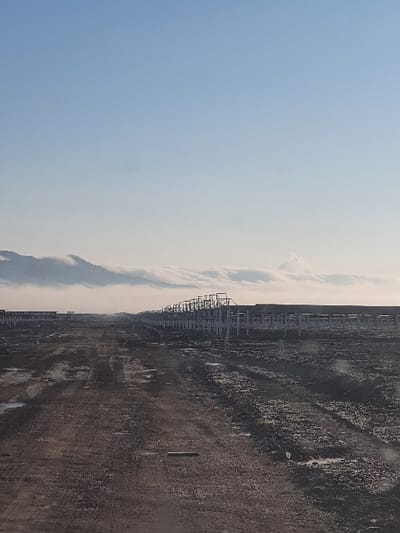 An expansive landscape unfolds under a gradient sky, transitioning from a clear blue to a pale, cloud-streaked horizon. Along the dividing line of sky and earth, a structure, possibly industrial, creates a repetitive, linear pattern. The ground is predominantly a rugged terrain of dark soil and debris, with subtle hints of moisture reflecting light. The overall scene presents a stark contrast between the natural sky and the human-altered land.