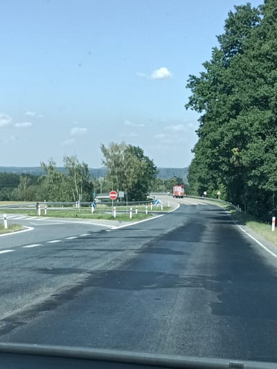 This image captures a view from a vehicle driving on a paved road in a rural, possibly forested, setting. The road is wet in patches, suggesting recent rain or morning dew. White lane markings are visible, indicating the road is a two-lane highway.
The scene features a gentle curve in the road, with a large, dense green tree line dominating the right side of the frame. On the left, a crossroads or intersection is present, marked by white bollards and a guardrail. Several traffic signs are visible at the intersection: a circular red and white "no entry" sign, a blue circular sign with a white arrow indicating a mandatory turn, and a green sign with a symbol that could be a directional sign.
In the distance, a red truck with a load is driving away from the camera on the curving road. The landscape beyond the road is characterized by rolling hills covered in green trees and vegetation, fading into a hazy horizon. The sky is a clear blue with a few scattered white clouds. The overall atmosphere is bright and natural, suggesting it is daytime, likely mid-morning or early afternoon, given the quality of light and the absence of strong shadows.
The location is identified as Šternov, Czechia, which aligns with the appearance of the landscape and the style of road signs. No visible text is present on the signs, apart from the symbols. The image conveys a sense of travel and the openness of a country road.