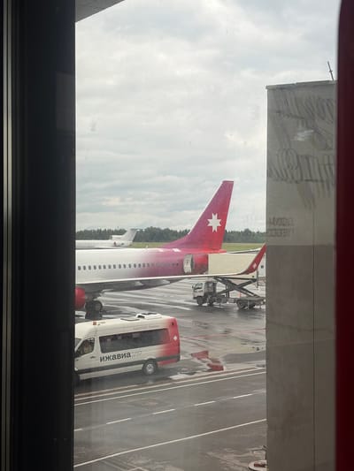 The image captures an airport tarmac scene viewed from an indoor window, with a dark frame in the foreground. The ground is wet, suggesting recent rain.
The central subject is the tail section of a passenger aircraft, oriented with its nose pointing right. The aircraft's vertical stabilizer (tail fin) is a distinctive red/maroon color, adorned with a prominent white, multi-pointed star emblem, characteristic of Izhavia airline. The fuselage is primarily white, transitioning to a subtle gradient or pattern of red/purple near the tail. Ground support equipment, including a baggage loader, is positioned next to the aircraft, indicating ongoing or impending ground operations.
To the right of the aircraft, a white and red airport shuttle bus is parked. Visible text on its side clearly reads "ИЖАВИА.VIP" and "Аэропорт Ижевск" (Izhevsk Airport), confirming the location as Izhevsk, Russia.
The sky is overcast, contributing to the muted lighting of the scene. The wet asphalt reflects the sky and the surrounding environment. Faint, mirrored text and patterns are visible as reflections on the window glass in the upper section of the image, partially obscuring the view of the sky and a building's roofline. The overall atmosphere depicts a typical daytime airport ground operation under cloudy weather conditions.