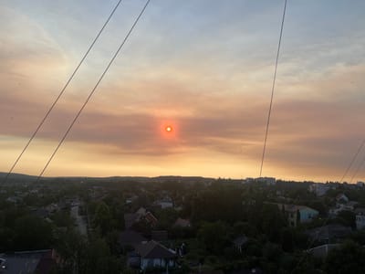This is a wide-angle, high-angle view of a suburban area in Vasylkiv, Ukraine, during sunset. The sky is a gradient of soft blues and oranges, with a hazy orange glow emanating from the sun, which is partially obscured by atmospheric conditions. Several power lines are visible in the foreground, stretching diagonally across the frame. Below the sky, a dense arrangement of residential buildings, interspersed with numerous trees, stretches across the landscape. The rooftops vary in color and material, with some appearing tiled and others with darker surfaces. A winding road is visible in the mid-ground, with no discernible vehicles or pedestrians. The overall impression is of a quiet, rural or suburban setting during dusk.