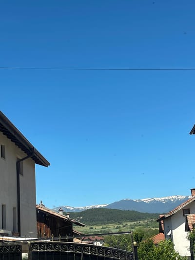 The image depicts a view towards snow-capped mountains from what appears to be a residential area in Kraishte, Bulgaria. A clear blue sky occupies the majority of the frame. Architectural elements, including rooftops with brown tiles, a white building with windows, and a wrought-iron fence, frame the view. The landscape transitions from the buildings to a green hillside leading up to the snow-covered peaks. A power line is also visible against the sky.