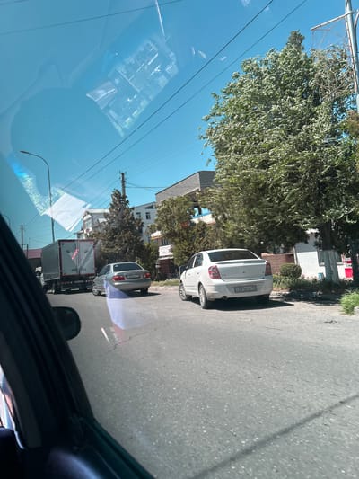 The image shows a street scene in Аксукент, Kazakhstan, taken from inside a vehicle. The view includes two parked cars on the right side of the street: a white hatchback and a silver sedan. Further down the street, a truck with a white cargo container is visible. Trees line the street, and buildings are seen in the background. Overhead electrical wires stretch across the sky. The blue sky is reflected in the windshield of the vehicle taking the picture, and parts of the car's interior are also visible.