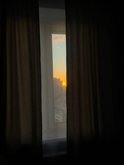 An interior shot captures a vertical segment of a window in Dachnoe, Russia, framed by two dark, heavy curtains drawn mostly closed. The curtains, dark brown with discernible vertical pleats and texture, occupy the majority of the left and right sides of the image. A white window frame is visible in the central opening. Through this aperture, the sky at what appears to be sunrise or sunset is visible, presenting a light blue hue with subtle orange and pink cloud formations in the upper section. Below the sky, silhouettes of multiple distant buildings are apparent, partially obscured by atmospheric effects. A bright, intense orange light source, presumed to be the sun, is positioned slightly to the right of the vertical center within the visible window area, casting a strong glow. Beneath the distant structures, a wide, reflective surface, possibly a body of water or a road, is present, displaying faint patterns indicative of ripples or textures. The lowest visible part of the exterior view is partially obscured by a dark interior element, potentially a windowsill. No individuals, animals, or explicit actions are observed.