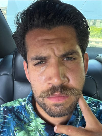 The image is a close-up, sideways portrait of a man sitting in a car. He has dark hair, dark eyebrows, and a light beard with some blonde hairs mixed in. His eyes are dark. He is wearing a blue and green floral print shirt. His right hand is raised, with his index finger touching his cheekbone. The background shows the interior of a car, with black leather seats visible. Through the window, there are reflections of buildings and greenery, suggesting an urban or suburban outdoor setting. The lighting suggests it is daytime. The man appears relaxed. No visible text is present in the image.